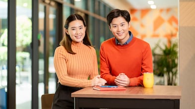 Two people stand at a high table in a modern office space, with a tablet placed on the surface and a yellow device nearby, against a bright background with large windows and decorative wall patterns.