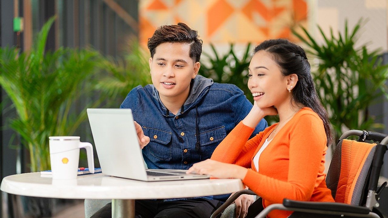 Two people sitting at a round table, looking at a laptop screen, with a coffee mug and plants in the background.