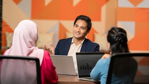 Three people seated at a table with laptops, engaged in a discussion in a modern office with geometric wall design.