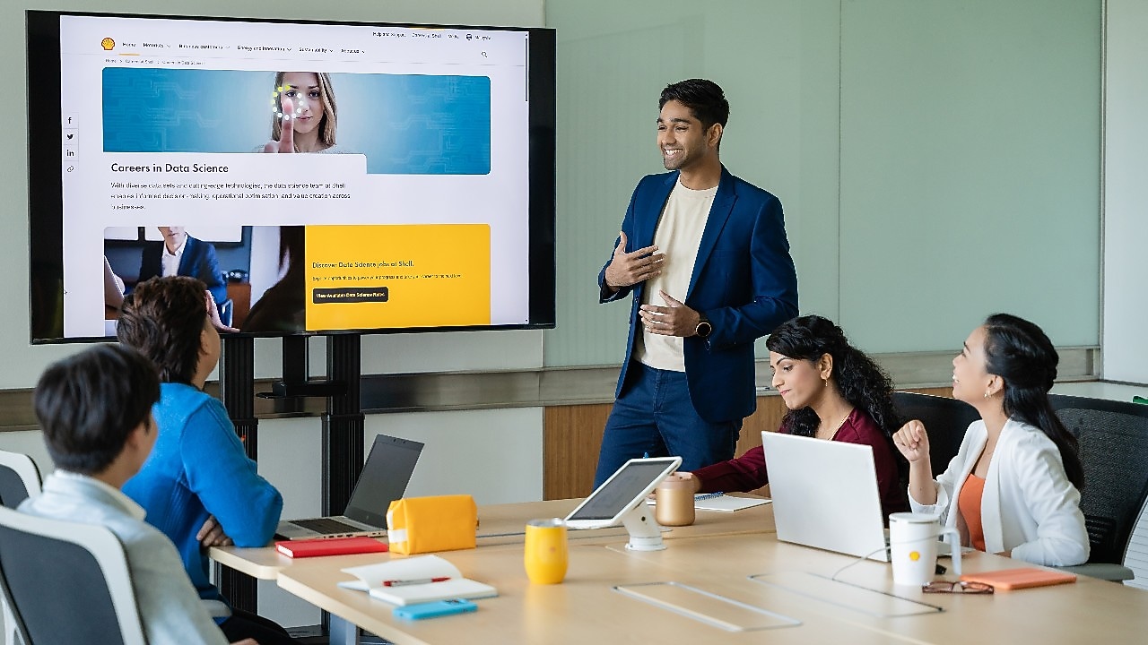 Person presenting a data science careers webpage on a large screen to a group seated at a conference table with laptops.