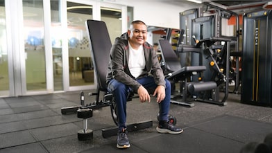 A person sits on a weight bench in a gym, wearing a gray hoodie, white t-shirt, blue pants, and sneakers. The gym background features various exercise machines and weights.