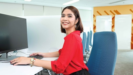 person in a red shirt with short hair is seated at a white desk in a modern office, working on a computer