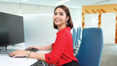 A person in a red shirt with short hair is seated at a white desk in a modern office, working on a computer.