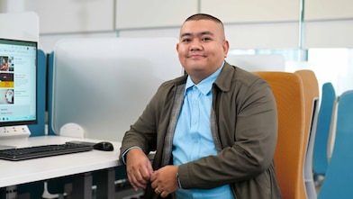 person seated in a modern office environment, wearing a light blue shirt and gray jacket, sitting on an orange chair. In front of them is a desk with a computer monitor displaying a webpage, along with a keyboard and mouse