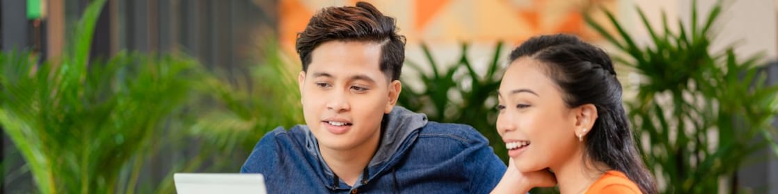 Two people sitting at a round table, looking at a laptop screen, with a coffee mug and plants in the background.