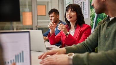 People seated at a conference table with laptops, discussing data shown on charts and graphs in a meeting room.