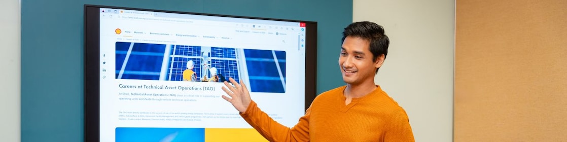 Person giving a presentation in a meeting room, pointing at a large screen displaying a webpage, with two colleagues seated.