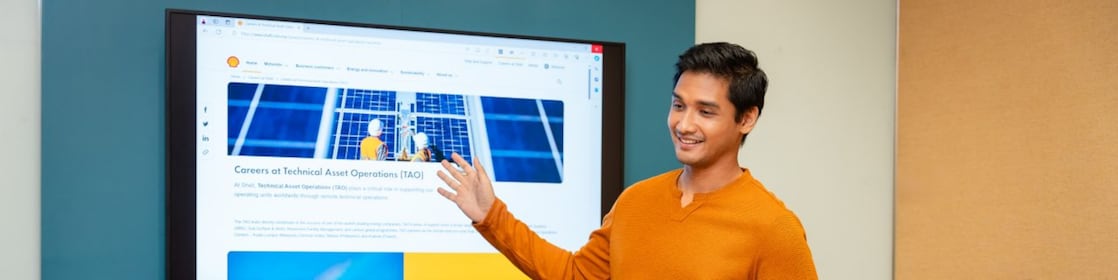 Person giving a presentation in a meeting room, pointing at a large screen displaying a webpage, with two colleagues seated.