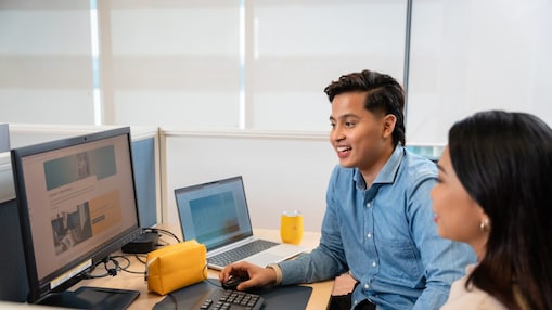 Two people sit at a workstation in an office, reviewing content on a desktop monitor and laptop, with a yellow pouch and a glass of orange drink on the desk.