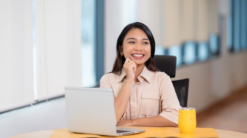 A person sits at a desk in a bright office, working on a laptop. A glass of orange-colored drink is placed on the table beside the computer, with large windows and a blurred workspace in the background.