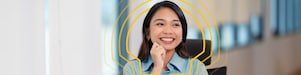 A brown-skinned woman seated at a desk with a laptop and a yellow cup, smiling and resting her chin on her hand. Background shows a blurred office space.