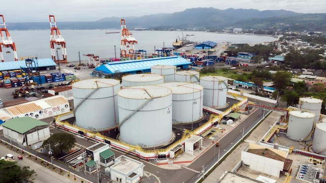 Aerial view of a fuel storage terminal beside a busy seaport, with large storage tanks, cargo cranes, container yards, and a coastal mountain backdrop.