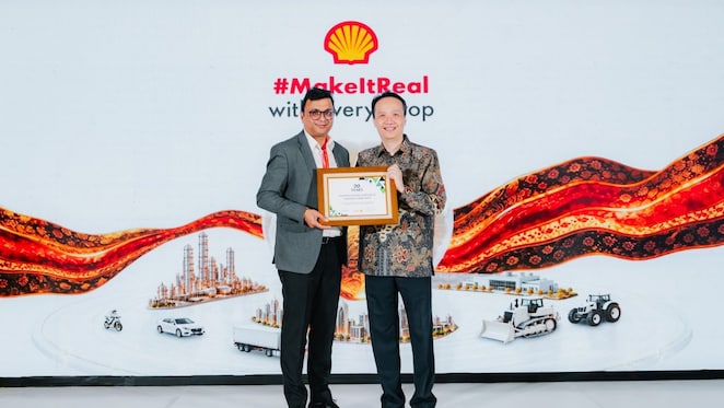 Two individuals stand in front of a Shell-branded event backdrop, jointly holding a framed recognition certificate. The backdrop features the Shell logo, the text “#MakeItReal with every drop,” and illustrations of industrial facilities, transportation vehicles, and agricultural machinery connected by a flowing red and orange graphic.