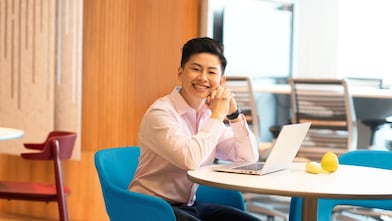Person with short dark hair, dressed in a light pink shirt, smiling confidently while sitting at a white table with a laptop, banana and a lemon, in a colorful, modern office background.