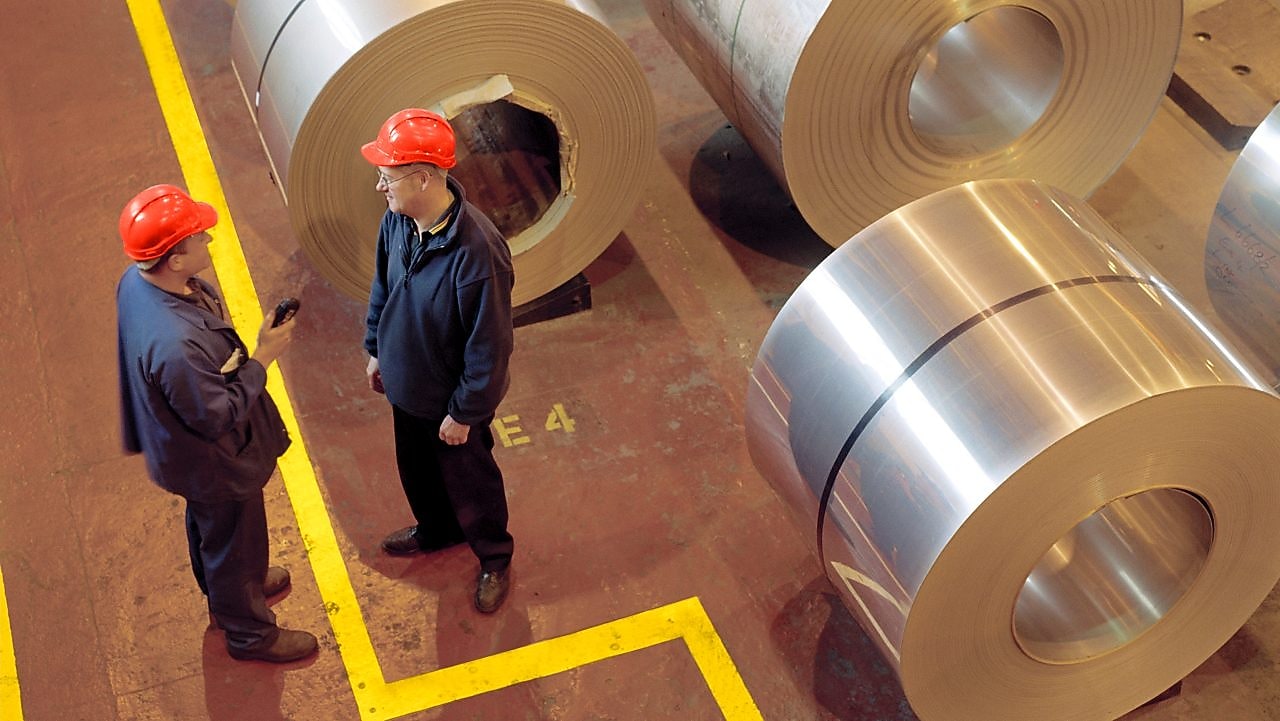 Two factory workers standing next to rolls of steel