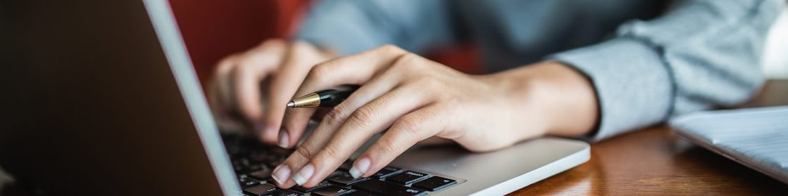 Person typing on a laptop while holding a pen, working at a wooden desk with a notebook nearby.